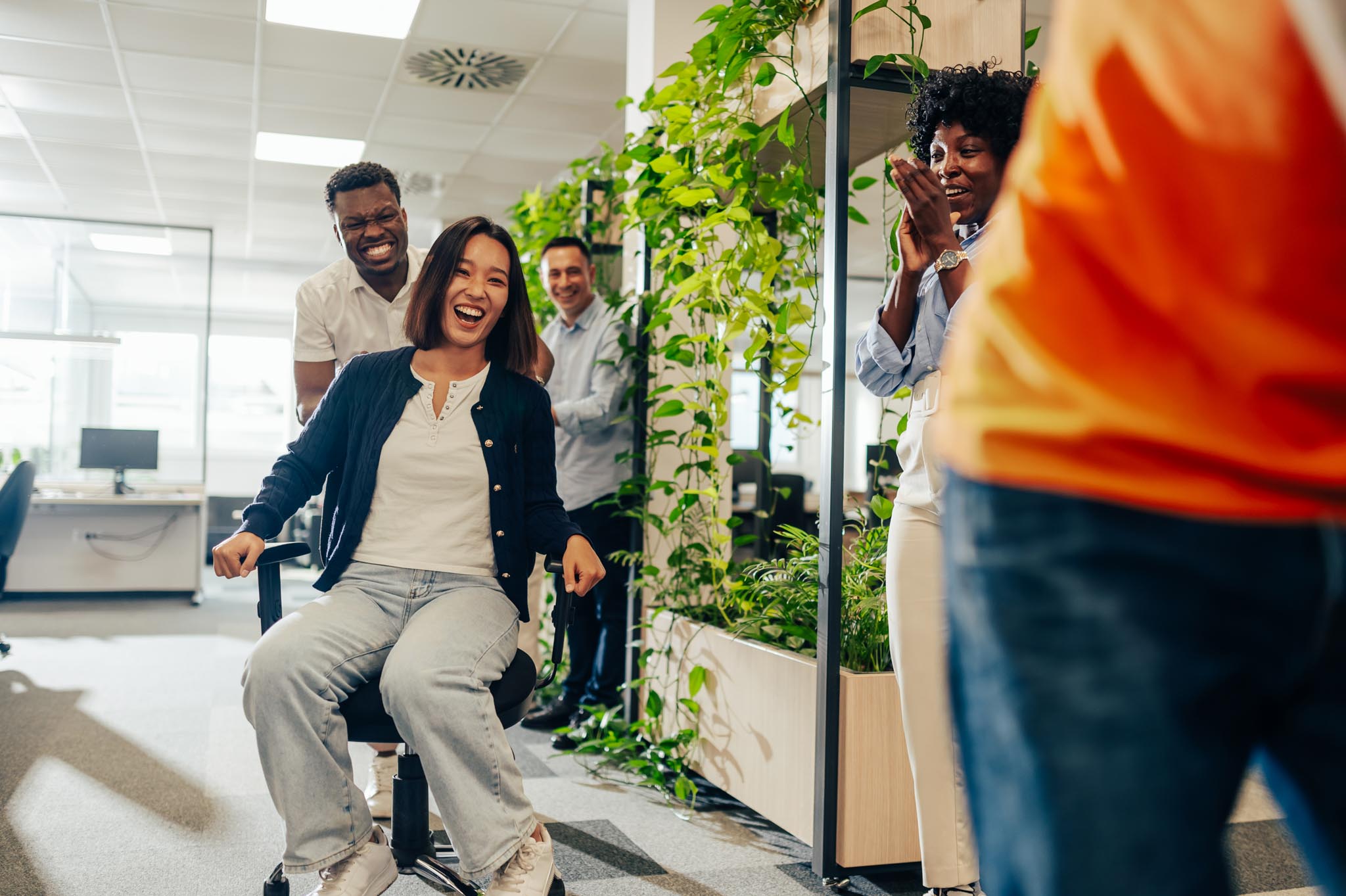 Diverse people having fun at work and racing on office chairs. Asian chinese businesswoman sitting on a chair while her african american man coworker is pushing her and other are cheering. Copy space.