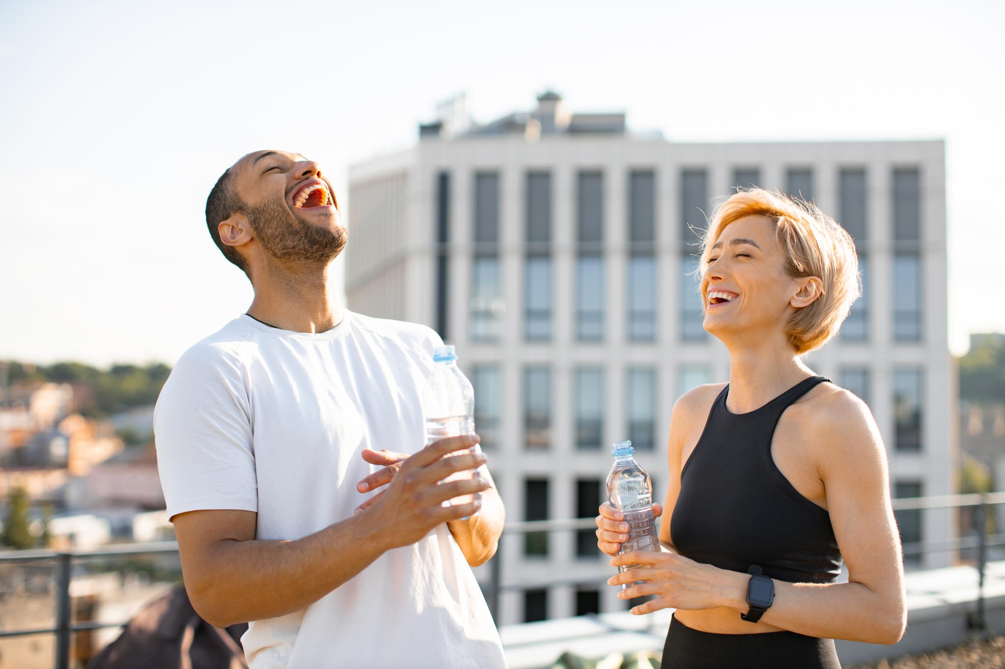 Young couple having fun laughing after morning workout on the roof of the house. Athletic man in white t-shirt and woman in black top drinking water after jogging.